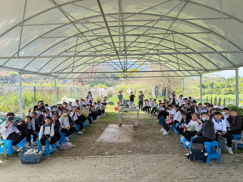 A big group listening to a talk under a farm shelter